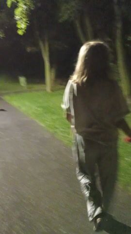 The video shows a person walking on a paved path at night. The path appears to be in a park or wooded area, with grass and trees visible. The person is wearing a light-colored t-shirt and dark pants. The lighting is dim, suggesting it is nighttime. The person is walking away from the camera, and their face is not clearly visible. There is another person in the background, also walking. The overall mood is casual and relaxed, with no indication of any particular event or activity beyond walking. The location is identified as Sokolov, Czechia.