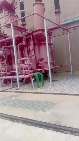 The video pans across an outdoor industrial site, likely a power generation facility. In the foreground, there are grates and a concrete walkway with tracks. The primary focus is a large, red industrial transformer with numerous pipes and gauges. Further in the background, there are more transformers, concrete buildings, and a large, rocky hillside. The scene appears to be a functional, operational area within a large power station, possibly a hydroelectric dam given the mountainous backdrop. There are no people visible in the video.
