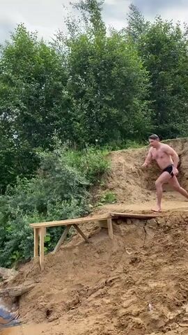 A man in black swim trunks stands on a wooden plank platform, poised to jump. Behind him, a dense green forest and a sandy embankment form the background. He leaps forward, his body arcing through the air. He plunges into the clear water, creating a large splash. The camera captures his descent into the water and the subsequent ripples on the surface. He then emerges from the water and begins to swim. The environment appears to be a natural lake with a sandy shore and a forested hillside. The sky is partly cloudy.
