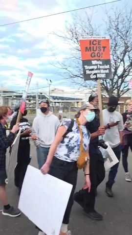 The video shows a group of people protesting in what appears to be an urban or suburban area. The protesters are holding signs with messages such as "ICE MUST GO! Whole Trump Fascist Regime Must Go NOW!" and "QUE SE LARQUE TRUMP! YA!". Some people are wearing masks or face coverings. The environment includes trees, buildings, cars and street fixtures such as traffic cones. The protest appears to be against the Trump administration's policies, particularly those related to immigration enforcement (ICE). The video does not capture the exact location or the overall size of the protest.
