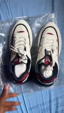 The video shows a pair of new, off-white sneakers with red and navy blue accents being presented on a light blue surface, possibly a bed sheet. The sneakers are sitting on a clear plastic bag with manufacturing information printed on it. A hand with manicured nails and a ring is rotating and adjusting one of the shoes to show its different angles, including the sole, the sides, and the interior, focusing on details like the branding inside the shoe. The video appears to be a product showcase, highlighting the design and features of the sneakers.