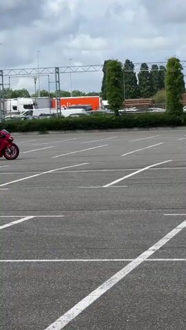 A motorcycle rider in a red and white suit is practicing on a red motorcycle in an empty parking lot. The rider is maneuvering around orange cones. There are other motorcycles and people in the background. The weather appears to be overcast.