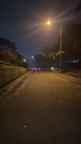 The video begins with a view of a road at night, illuminated by a streetlamp. A car with red taillights is visible in the distance. As the video progresses, the perspective shifts to a moving vehicle, likely a car, as it travels down the road. Palm trees and other foliage line the sides of the road. The road surface appears wet, suggesting recent rain. The sky is dark, with some faint lights visible in the distance. The motion blur increases, indicating the vehicle is accelerating. The video ends with a fast-moving shot of the road at night.