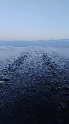 The video shows a flat body of water under a clear blue sky, with a narrow band of light clouds on the horizon. The dark water is disturbed by ripples and a central wake, suggesting a boat or other vessel is moving away from the viewer. The scene is quiet and still, save for the water's surface disturbance.