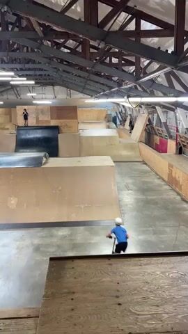 This video shows a person on a scooter at an indoor skate park. The skate park is made of wood and has several ramps and obstacles. The person is wearing a helmet and knee pads. The person is riding the scooter down a ramp and then performs a trick. The video is shot from a high angle, looking down at the skate park. The lighting is artificial, with fluorescent lights on the ceiling. There are other people in the background, some watching and some on other equipment. The person says "For the vlog, for the vlog."