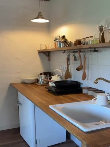 The camera pans slowly across a cozy kitchen counter.  The counter, made of warm-toned wood, holds a white porcelain sink filled with clean water. Beside the sink, a decorative white mug sits, and a vintage-style brass faucet stands tall.  To the left of the sink, several small ceramic jars with wooden lids are neatly arranged.  Further left, a black pan rests on a portable electric burner next to two silver kettles.  Wooden cooking utensils hang above the counter on a metal rack. Above the rack, a wooden shelf displays various kitchen items including glassware, utensils, and small decorative objects.  The wall behind the counter is covered in patterned white tiles, and a small white refrigerator is visible below the counter on the left.  A ceiling light illuminates the scene with a warm glow.  Background music plays.