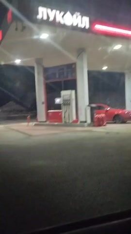 This media file captures a nighttime scene at a Lukoil gas station in Zelenchukskaya, Russia. The primary object of focus is a red car at one of the gas pumps. The gas station is illuminated by overhead lights and the prominent Lukoil logo, which is clearly visible at the top of the structure. The scene appears to be at night, with the dark sky and artificial lighting creating a somewhat stark atmosphere. The weather is not explicitly discernible from the image, but the presence of lights suggests it is not heavily raining or foggy. The activity taking place is refueling a vehicle, as indicated by the car positioned at the pump and the presence of gas hoses. Other vehicles can be seen in the distance on the road, suggesting continued traffic flow. There are no people visibly present in the immediate vicinity of the car or pumps, adding to a sense of quiet efficiency in the operation. The overall impression is a functional, albeit somewhat isolated, moment at a service station during the late hours.
