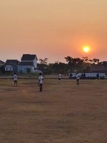 The video shows an outdoor scene at sunset. The sky is a warm orange and yellow as the sun is low on the horizon. In the foreground, a dry, grassy field is visible. Several people, likely young men, are scattered across the field, some wearing athletic clothing. They appear to be playing or practicing a sport, possibly soccer, as some are in a stance that suggests they are waiting for or interacting with a ball. In the background, there are some houses and trees. The overall atmosphere is peaceful and suggests an evening recreational activity.