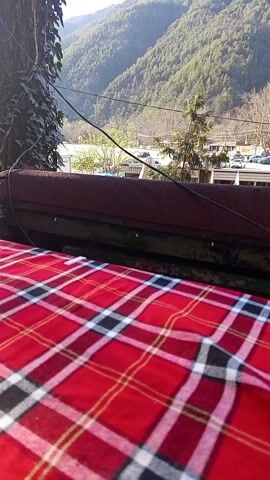 Here's a detailed description of the video:

The video presents a static view of a rustic outdoor setting, likely a cafe or restaurant with a scenic mountain backdrop. The foreground is dominated by a table covered with a red plaid tablecloth. Above the table, a weathered wooden structure is visible, partly obscured by a tree trunk covered in ivy. A vintage Coca-Cola sign hangs from the structure above the table. 

In the background, a lush green mountain range extends across the horizon, adding a sense of depth and tranquility to the scene. Below the mountains, a small town or village is visible, with buildings, cars, and trees. 

The lighting suggests a bright, sunny day, enhancing the colors of the tablecloth and the natural beauty of the mountains. Overall, the video captures a peaceful and charming scene, blending natural elements with a touch of vintage aesthetics.