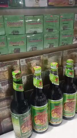 A video shows a shelf display in what appears to be a shop or market. The shelves are stocked with various products, primarily in green and brown packaging. In the foreground, a row of dark glass bottles with green and yellow labels is arranged on a counter. The labels on these bottles depict a honeycomb pattern and text that appears to be in a non-English language, possibly Indonesian. Behind the bottles, more products are visible on shelves, including boxes with similar green and brown designs. The lighting suggests an indoor setting. There are no people visible in the video. The camera pans across the display, offering a closer look at the bottles and the products on the shelves.