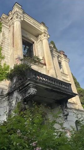The video shows a low-angle shot of an abandoned, decaying building with a classical architectural style. The building has columns, a balcony, and multiple floors with empty window frames. The facade is weathered and overgrown with greenery, including vines and bushes. The sky is visible through the window openings. The camera slowly pans upwards, revealing more of the building's height and the extent of its decay.