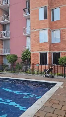 The video shows a rectangular swimming pool surrounded by a paved area in an outdoor setting. Several people, including adults and children, are in the pool. Some are swimming, others are standing or sitting in the water. One person is seen tossing a ball. A black fence runs along the perimeter of the pool area, separating it from the surrounding landscape. In the background, there are multiple apartment buildings with brick facades and balconies. A playground structure is visible near the pool. The video pans across the scene, capturing different angles of the pool and the people enjoying the water.
