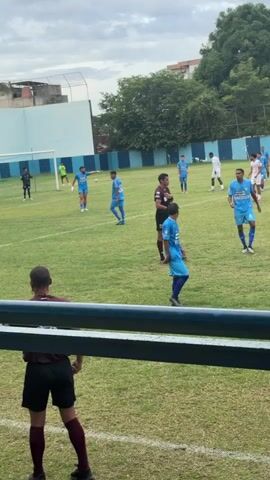 A referee in a burgundy shirt and black shorts pulls out a yellow card on a grass soccer field. Several soccer players in blue and white jerseys stand on the field around the referee. One player in a light blue shirt and dark blue shorts with the number 13 on his shorts stands closest to the foreground on the right. A light blue and white fence surrounds the soccer field. Trees and buildings are visible in the background.