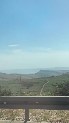 The video, taken from a moving vehicle, shows a landscape of rolling green hills under a clear blue sky. A body of water is visible in the distance, partially obscured by haze. The foreground features a grey highway guardrail, and along the road, there are intermittently placed light posts. A dirt path winds through the landscape. The overall impression is a sunny, rural scene.