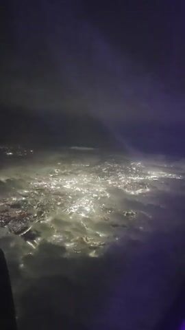 A nighttime aerial view from an airplane window shows a vast city illuminated by countless lights below, appearing as a sprawling network of glowing urban areas. Wisps of clouds or fog drift over the city, partially obscuring the lights and creating a hazy, ethereal effect. A portion of the airplane's wing or fuselage is visible in the bottom left corner of the frame. The overall scene is dark, with the city lights being the dominant source of illumination against the shadowy sky.