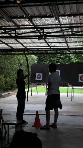 Two people are standing on a concrete floor under a corrugated metal roof. On the left, a person wearing dark clothing and a hat holds a bow, aiming it towards the right. They are standing next to a chair. To the right of this person is an orange traffic cone. Further to the right is another person wearing a white t-shirt and shorts, facing the person with the bow. Behind them are three archery targets mounted on stands. Lush green trees and foliage are visible in the background. The overall scene appears to be an outdoor archery range.