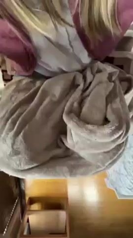The video shows a person sitting with a blanket on their lap. They lift the blanket to reveal a Sphynx cat curled up between their legs. The person is wearing blue jeans. The cat is hairless and appears pinkish. The scene is indoors with wooden flooring.
