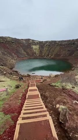 A high-angle shot shows a volcanic crater lake with a set of wooden stairs leading down towards it. The crater walls consist of red and brown rock formations, with some green vegetation. The lake itself has a greenish-blue hue. Several people are visible around the crater's edge and near the lake. The weather appears overcast. The stairs are constructed with wooden planks supported by side beams, creating steps down the slope.