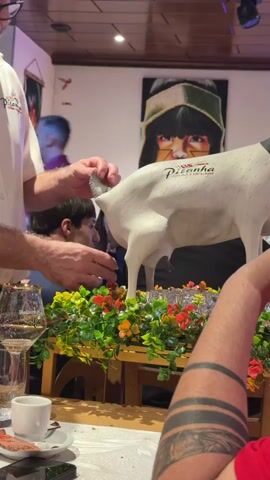 The video shows a close-up of a white, life-sized model of a cow on a table, adorned with artificial flowers and greenery. A man wearing a white shirt with a logo that reads "Picanha" is seen from the chest down, touching the model's tail. He is holding a knife in his left hand. In the background, another person is partially visible, and a painting of a person's face is hanging on the wall. The scene appears to be indoors, possibly in a restaurant, with a table set with glasses and a small cup. The lighting is warm, suggesting an evening or dimly lit environment.