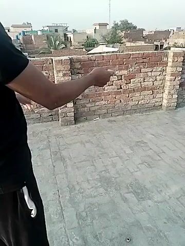 A person wearing a black t-shirt and black sweatpants with a white stripe stands on a rooftop next to a short brick wall. The person is holding a white string, likely kite string, in their hands, moving it back and forth. The rooftop is made of grey rectangular tiles. In the background is a view of a neighborhood with houses and a communication tower. The sky is overcast.