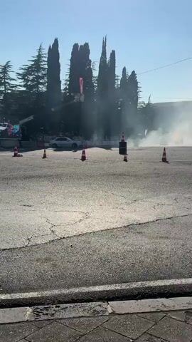 A white car is drifting on a paved road, creating a large amount of white smoke. There are several orange traffic cones placed on the road, marking a course. In the background, there is a crowd of people watching the event, and tall trees line the edge of the road. A traffic light and a red flag are visible above the crowd. The scene appears to be set outdoors during the daytime, with a clear sky. The overall atmosphere seems to be one of excitement and skill, as the driver maneuvers the car through the smoky drift. A black, rectangular object with orange markings is situated between two cones in the middle of the frame. The pavement is cracked and weathered. The video captures a moment of a drifting competition or demonstration. There is some visible text on a sign in the background, but it is too blurry to read. The lighting suggests it is a sunny day.