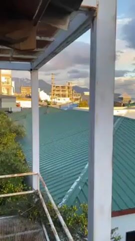 The video shows a view from a rooftop looking out over a town. The buildings are mostly low-rise, with a few taller structures in the distance. The roofs are mostly made of corrugated metal, some painted in different colors. The sky is cloudy with a hint of sunset. The camera pans across the scene slowly. There are metal structural elements in the foreground, suggesting that the view is from a building under construction or in need of repair.