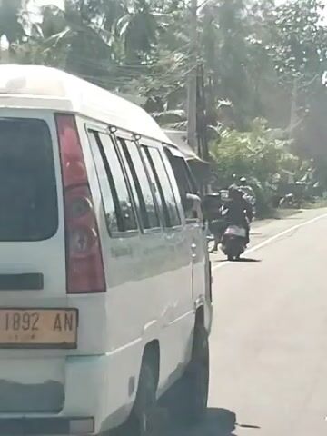 A white van with yellow license plate is parked on the side of a road. A person is riding a motorcycle and passes the van. Several cars are driving on the road in the distance. The road is paved and there are trees and vegetation on both sides of the road. The sky is blue and clear.