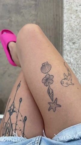 A static, high-angle shot displays a person's crossed legs clad in denim shorts. The individual wears bright pink sandals. The upper leg features several small, outlined tattoos: a scallop shell, a flower, a conch shell, a starfish, and a fish. The lower leg showcases a larger, more intricate tattoo. The background is a neutral gray, likely concrete or tile.