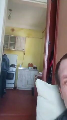 The video shows a person resting against a white pillow, possibly in a room, with a view into a small kitchen. In the kitchen, a younger person in a dark jacket stands near a stove, where a pot sits. The kitchen walls are a pale yellow color, and there are cabinets above the counter. The room is illuminated by natural light. The scene appears to be indoors, possibly in a modest or older dwelling. The resting person is close to the camera, focusing the viewer's attention on their face while the kitchen scene serves as a background.
