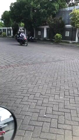 The image depicts an outdoor scene on a paved street. The foreground is dominated by a textured brick-patterned surface, suggesting a walkway or road. In the mid-ground, two individuals are riding a motorcycle, both wearing helmets. The rider is in a white shirt and the passenger is in a dark shirt. Behind them, another person is seen standing next to a parked motorcycle. Further in the background, there's a building with a fence and lush greenery, including trees and bushes. The scene appears to be set during the daytime, with natural lighting. The overall mood is casual and ordinary, with no strong emotional indicators. There is no visible text in the image. The location context provided is Boro, East Java.
