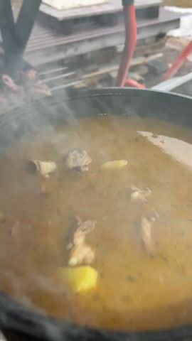 The video captures an outdoor cooking scene in Bol'shakino, Russia, likely during cold weather given the visible snow on the ground. A large, dark-colored pot is filled with a simmering, yellowish-brown stew or soup, from which a significant amount of steam rises. Inside the pot, chunks of meat and pieces of potato are visible.

Adjacent to the pot, a metal grill is in use, with skewers of meat cooking over hot coals. Smoke and steam emanate from both the grill and the pot, creating a hazy atmosphere. The background shows an outdoor area with a wooden structure, possibly a fence or trellis, and patches of snow on the ground. The overall impression is one of hearty outdoor cooking. There is no visible text in the video.
