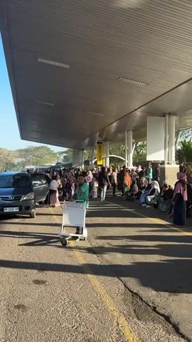 The video shows a crowded outdoor area, likely an airport or transportation hub, with a long covered walkway. In the foreground, a man in a green shirt with a badge is pushing a white luggage cart. He is looking towards the camera. Behind him, a large group of people are gathered, many of whom appear to be waiting. Some are standing, while others are sitting on benches or the ground. The people are dressed in a variety of casual clothing, with many women wearing headscarves. Cars are visible on the left side of the frame, and a yellow sign with the words "DROP ZONE" is hanging from the covered walkway. The overall impression is one of a busy and possibly disorganized waiting area. The lighting suggests it is daytime, with sunlight casting shadows on the ground.