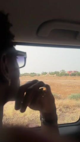 The video shows a person's hand and arm resting on a car window as they look out at a dry, grassy landscape. The person is wearing sunglasses, a watch, and a gold ring. In the distance, there are some buildings and trees. The sky is overcast.