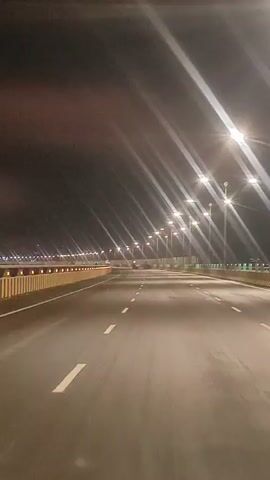 A dashcam footage captures a view of an empty highway at night. The highway is illuminated by streetlights which create bright streaks of light. The road has white lane markings. On the right side of the road, there is a yellow and white striped barrier. The sky is dark and appears to have some clouds or haze. The footage shows movement, suggesting the vehicle is in motion.