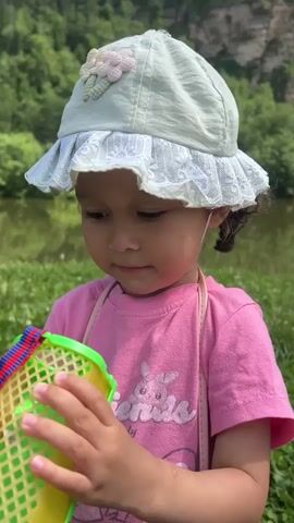 A close-up shot shows a young girl with dark curly hair wearing a light green hat with decorative flowers and a pink t-shirt with a graphic design. She is holding a colorful plastic container with a mesh exterior, possibly for catching insects, and appears to be looking at something inside it. The background suggests an outdoor environment with greenery and a body of water.