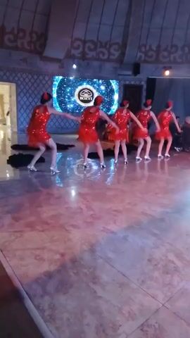 A group of five young women in matching red sequined dresses and red headbands are performing a dance routine on a polished floor in a dimly lit room. The women are positioned from left to right, spaced evenly, and are all facing forward. They appear to be in sync, with their arms raised and bent at the elbow, and their bodies slightly angled. Behind them, a large screen displays a circular, starry pattern with a logo in the center. The room has a high, vaulted ceiling with decorative patterns and is lit by ambient lighting and a bright spotlight. To the far left, a doorway is visible, and to the right, there are seated figures in the background. Black fabric lies on the floor in front of the dancers.