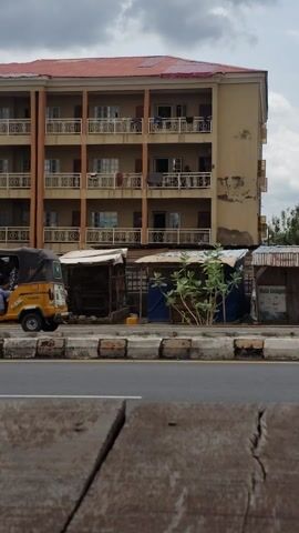 The video presents a street-level view, captured from a moving perspective, likely within a vehicle. The scene is dominated by a multi-story building on the left side of the frame, appearing to be residential with several floors of apartments or units. Each floor features balconies adorned with decorative railings, and clothes can be seen hanging to dry on some. The building's exterior is light brown or beige with a red-tiled roof.

Between the building and the asphalt road, several small, somewhat dilapidated structures or stalls are visible, constructed from materials like corrugated metal and wood. These structures suggest informal businesses or temporary shelters. A street light pole, equipped with a solar panel, stands near these stalls.

The road itself is paved with asphalt, bordered by a curb painted with distinctive black and white stripes. The sky above is overcast, indicating a cloudy daytime setting. To the right of the road, a textured surface, possibly a wooden panel or part of the vehicle's interior, partially obscures the view. Scattered greenery and trees are also present throughout the scene. No people or discernible text are visible. The overall impression is that of an urban or semi-urban environment in Adankolo, Nigeria.