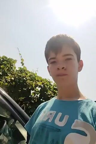 The video begins with a close-up of a young person's face, looking towards the camera with a neutral expression. The person is wearing a blue t-shirt. The camera then pans to show the person reaching for the door handle of a black car. The background shows green foliage and a bright sky. The person opens the car door and gets into the car. The interior of the car is visible, and the person is looking directly at the camera with a surprised expression. The video ends abruptly.
