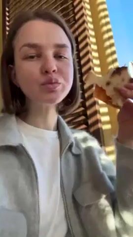 The video shows a young woman sitting indoors, eating a piece of pizza-like flatbread. She is wearing a white t-shirt and a light gray fleece jacket. She looks directly at the camera while chewing, then looks slightly to the side. The background features blinds with sunlight shining through, creating shadows. The sky outside the window is a clear blue.