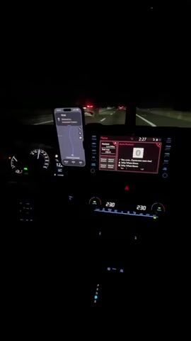 The video shows the interior of a car at night. The car is moving on a highway, as indicated by the road markings and other cars visible ahead. The dashboard is illuminated, with a navigation app displayed on a phone mounted to the windshield. The car's infotainment system is also visible, showing music playing. The driver's side dashboard shows the speedometer, which indicates the car is traveling at approximately 120 kilometers per hour. The temperature control is set to 23 degrees Celsius. The overall ambiance is dark, with the interior lights and the glow from the screens providing illumination. The music playing is a pop song with Russian lyrics.