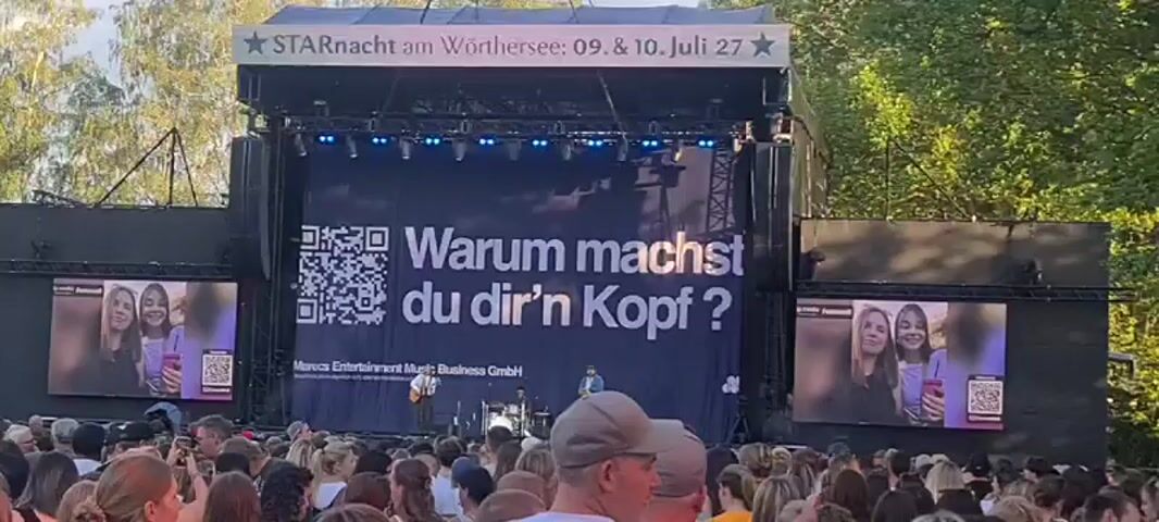 The video shows a concert stage with a large black backdrop displaying a QR code and the German text "Warum machst du dir'n Kopf?". The text translates to "Why are you worrying?". Two large screens on either side of the stage show images of two young women holding phones. A live band is performing on stage, with a singer in the center. A large crowd of people is gathered in front of the stage, many holding up their phones. The event appears to be an outdoor concert.