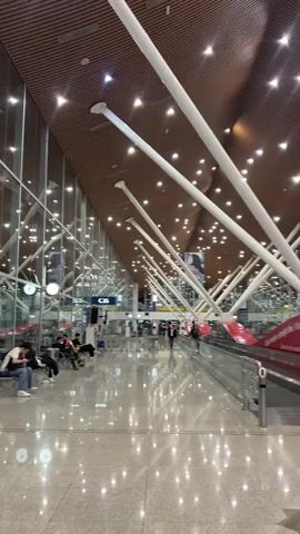 The video shows the interior of a spacious, modern airport terminal, likely Kuala Lumpur International Airport in Sepang, Malaysia, given the architectural style. The scene is brightly lit, with numerous small, recessed lights illuminating a high, wooden slatted ceiling. Large, white, slanted support beams extend from the floor or lower wall sections upwards towards the ceiling, creating a distinctive architectural feature.

The floor is a highly polished, reflective surface, mirroring the overhead lights and structural elements. On the left side of the frame, large glass panels form the outer wall, offering glimpses of what appears to be another section of the airport or exterior.

Several people are visible in the terminal. Some are seated on benches to the left, appearing to wait, while others are walking, including a few figures further down the concourse. A moving walkway, with distinctive red side panels, occupies the right side of the frame, running parallel to the main walking path.

Notable details include a gate sign displaying "C35" prominently in the middle-left of the shot. A large red banner or advertisement is visible on the right, above the moving walkway. Partially legible text on it reads "Life is rarely a straight line. Whe..." and "Connecting the world". The overall atmosphere appears calm, typical of an airport transit area, with a sense of order and modern design. The time of day is not explicitly clear from the natural light, but the artificial lighting makes the space very bright.