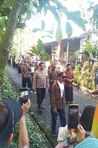 Here is a description of the video:

A group of people are standing in a line on a paved walkway. Most of them are wearing traditional striped shirts and headwear. One man in the center of the line is wearing a police uniform. Several people in the foreground are holding up cell phones, apparently filming or taking pictures of the group. In the background is a house with a red and white flag. The area is surrounded by trees and greenery. The camera is held vertically.