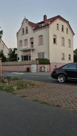 This media file depicts a street-level view of a multi-story building in Nierstein, Germany, likely during the late afternoon or early evening due to the soft, ambient light. The building is painted a light cream color with pale pink accents around the lower windows and base. It has a reddish-brown tiled roof and features multiple windows with beige shutters. A prominent feature is a white balcony with ornate railings on the first floor.

The setting appears to be a residential or mixed-use area with a road in the foreground and a sidewalk or pedestrian area with patterned paving stones. To the left of the building, a traffic light is visible, showing a green signal. A dark-colored SUV is captured in motion, driving past the building from left to right.

Other elements include a wooden fence with a pink top, some greenery, trash bins, and utility boxes. The sky is a pale blue, suggesting clear weather. A faint moon is visible in the sky above the building. The overall mood is calm and ordinary, showcasing a typical street scene. There is no discernible text visible in the file.