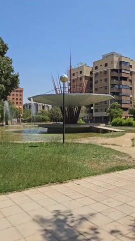This is a vertical video shot on a sunny day with a clear blue sky. The foreground shows a paved walkway with light brown square tiles, some of which are in shadow. To the left of the walkway is a patch of green grass. In the midground, there is a body of water with a fountain actively spraying water. A modern-looking sculpture with a large, umbrella-like canopy and thin, rod-like elements extending upwards is situated in the water. A single lamp post with a spherical light is positioned in front of the sculpture. Behind the water feature and sculpture are trees and several buildings, including apartment complexes of varying heights and architectural styles, appearing to be urban or suburban. The overall impression is a park or public square area.