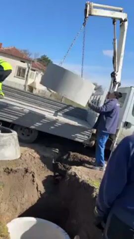 A crane lifts a concrete ring from the bed of a flatbed truck to place it into a hole in the ground. Three men are present at the scene. One man is operating the crane, another man is on the truck bed helping to guide the concrete ring, and the third man is standing near the hole. A concrete ring is already inside of the hole. The hole is dug into grassy land. A building can be seen in the background. The sky is blue.