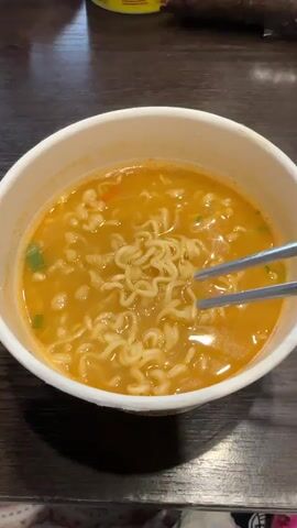Here's a description of the video:

The video shows a bowl of instant ramen being stirred with metal chopsticks. The ramen is in a white paper bowl filled with broth that is orange and slightly opaque. The noodles are curly and yellowish-white. There are visible green specks, possibly chopped scallions, and a few red pieces, likely dried chili. The chopsticks mix the noodles, then lift a portion of them to show their texture and shape. The bowl sits on a dark brown surface, possibly a wooden table, with a patterned item visible below the bowl. Another object sits out of focus in the background. The lighting appears to be indoor, likely artificial.