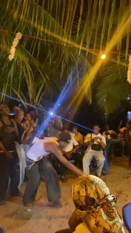 A group of people are gathered on a beach at night, watching and filming several individuals dancing in the center of the group. The dancers are moving energetically, and some of the seated people are clapping and cheering them on. A single blue spotlight is aimed towards the dancers. Palm trees provide shade, and strings of yellow lights illuminate the scene. The ground appears to be sand. The overall atmosphere seems festive and lively.