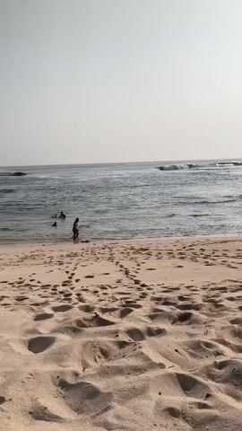 The video opens on a sandy beach with many footprints visible in the foreground. The ocean is in the background, with small waves rolling towards the shore. Several people are in the water. One person is walking from the beach into the ocean, and two other people are visible further out in the water. The sky is a pale, hazy gray. The video is taken from a low angle, looking out towards the water.