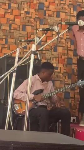 The video shows a young man seated and playing a bass guitar in what appears to be a church or event space. He is dressed in a light-colored, long-sleeved shirt and dark pants. A microphone stand is directly in front of him, holding several microphones. To his right, a person is standing in a pink shirt and dark pants. A large speaker is positioned behind the musician. The background consists of a wall made of multicolored brick or tile. The musician briefly puts his hand to his forehead before continuing to play. A voice is heard saying "Praise God."